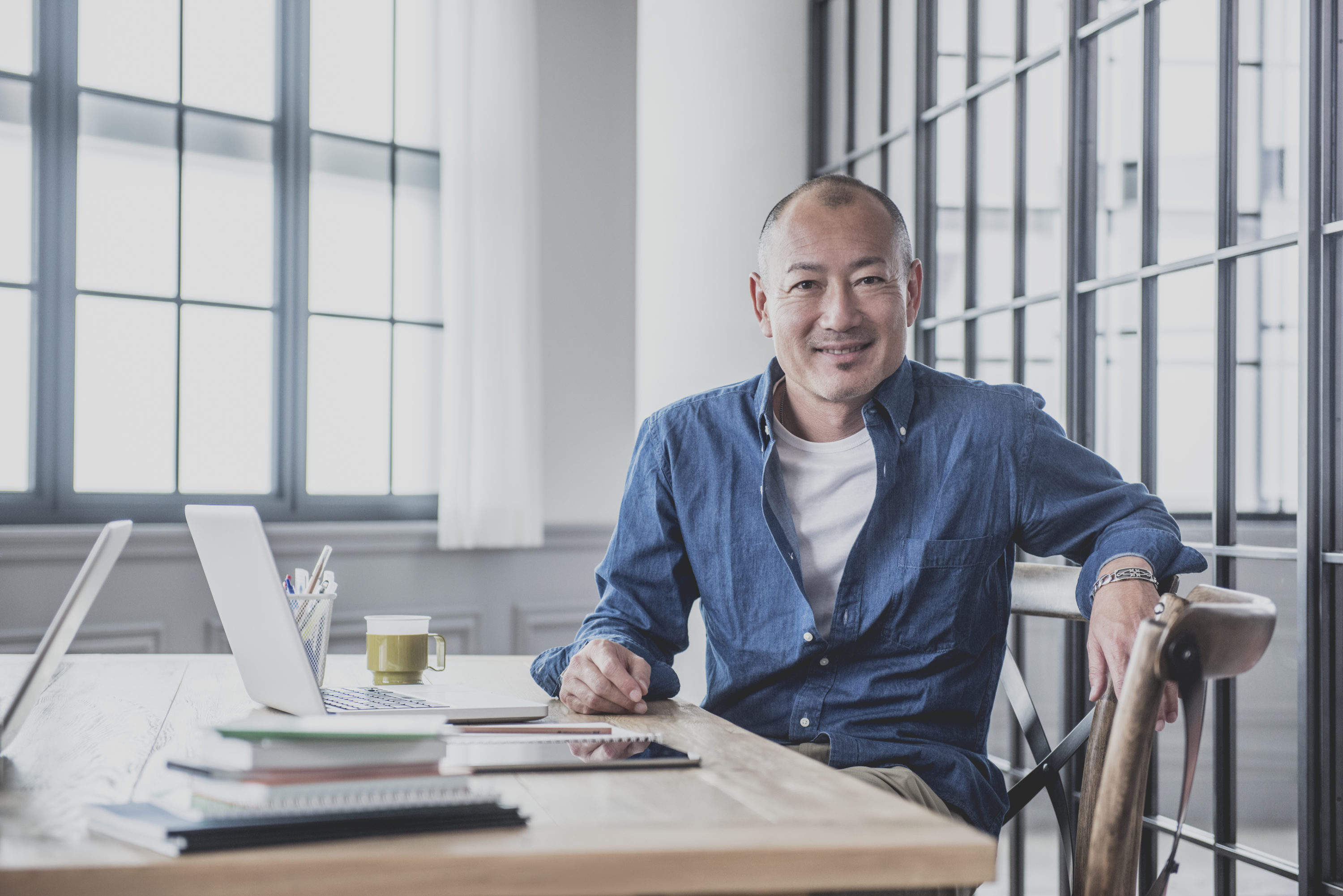 Japanese professional male worker sitting in modern studio with laptop, smiling to camera, Tokyo, Japan. Candid portrait of Asian man at work in creative agency.