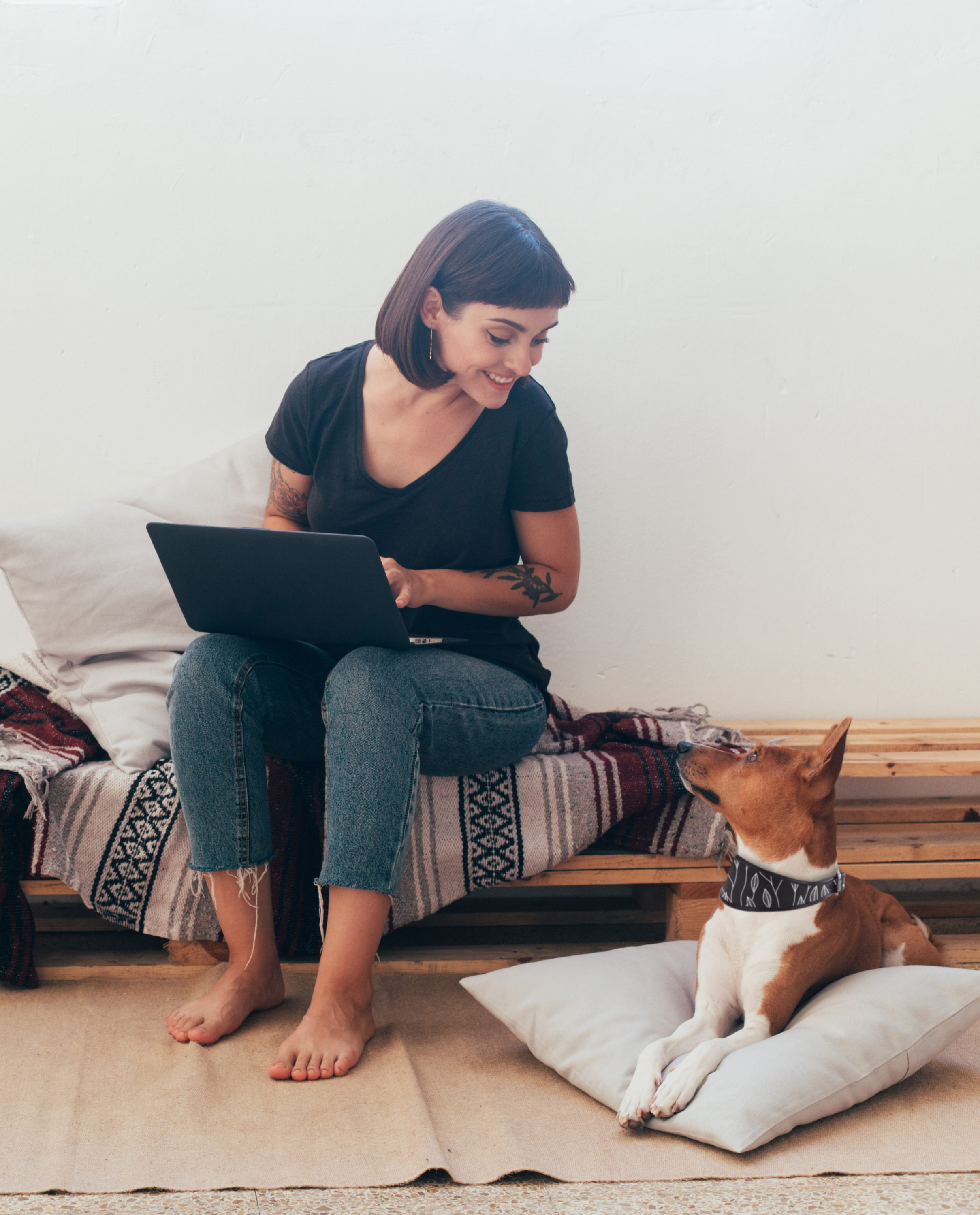 Pretty woman works from home or startup coworking space, sits barefoot on bench and writes code or blog on laptop, her best friend dog puppy lays next to her on pillow; Shutterstock ID 745832476; Purchase Order: -